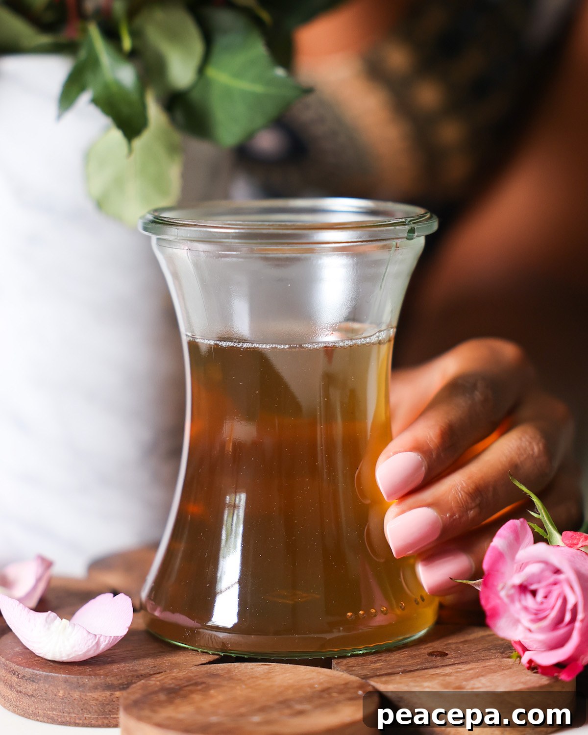 rose simple syrup in a glass jar held by a brown skinned hand, surrounded by rose petals.