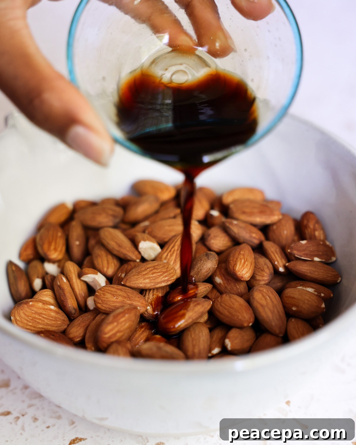 Drizzling a small dish of tamari over hot roasted almonds in a white bowl.