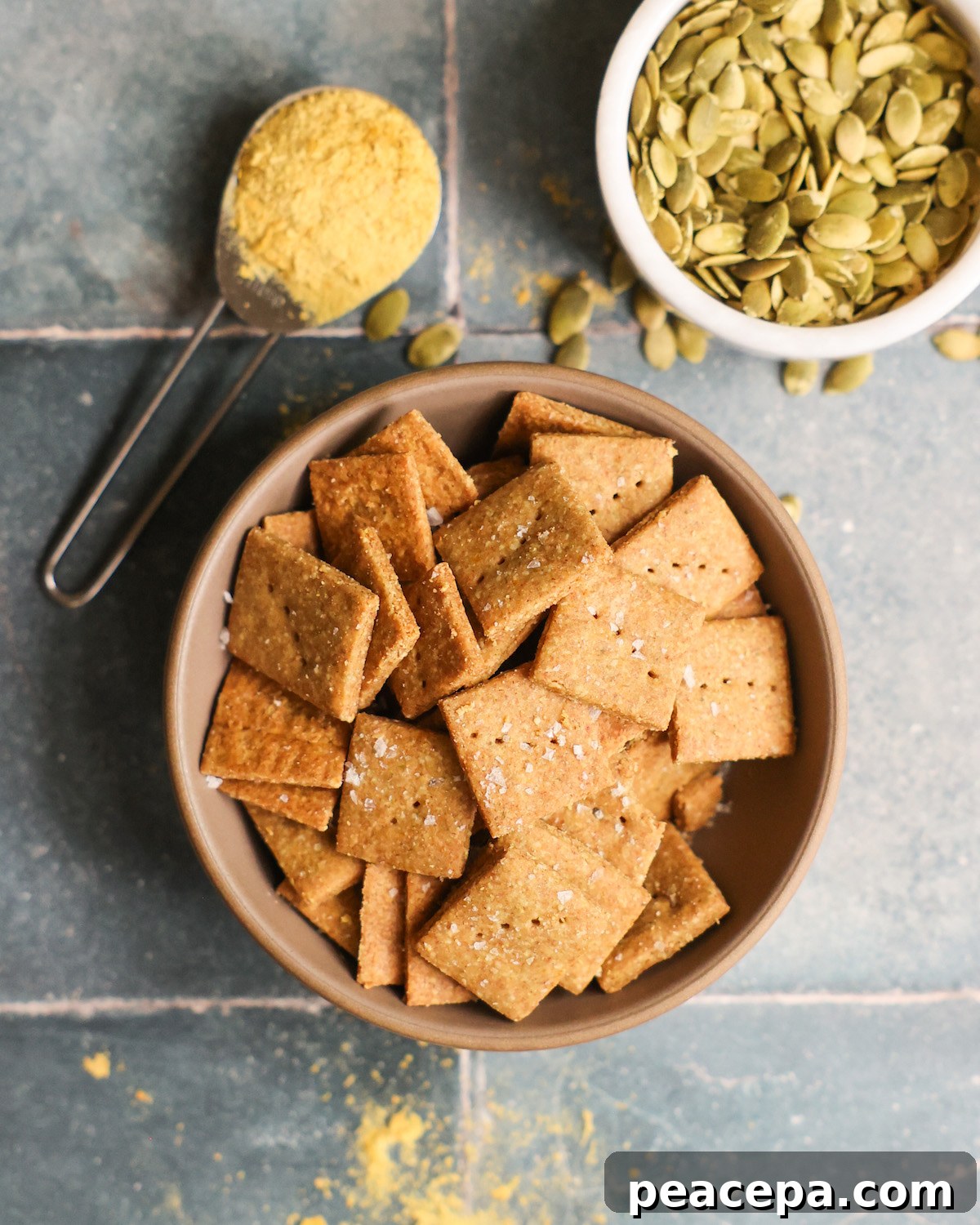An overhead shot of a bowl of pumpkin seed crackers on the counter with pumpkin seeds and a spoonful of nutritional yeast.