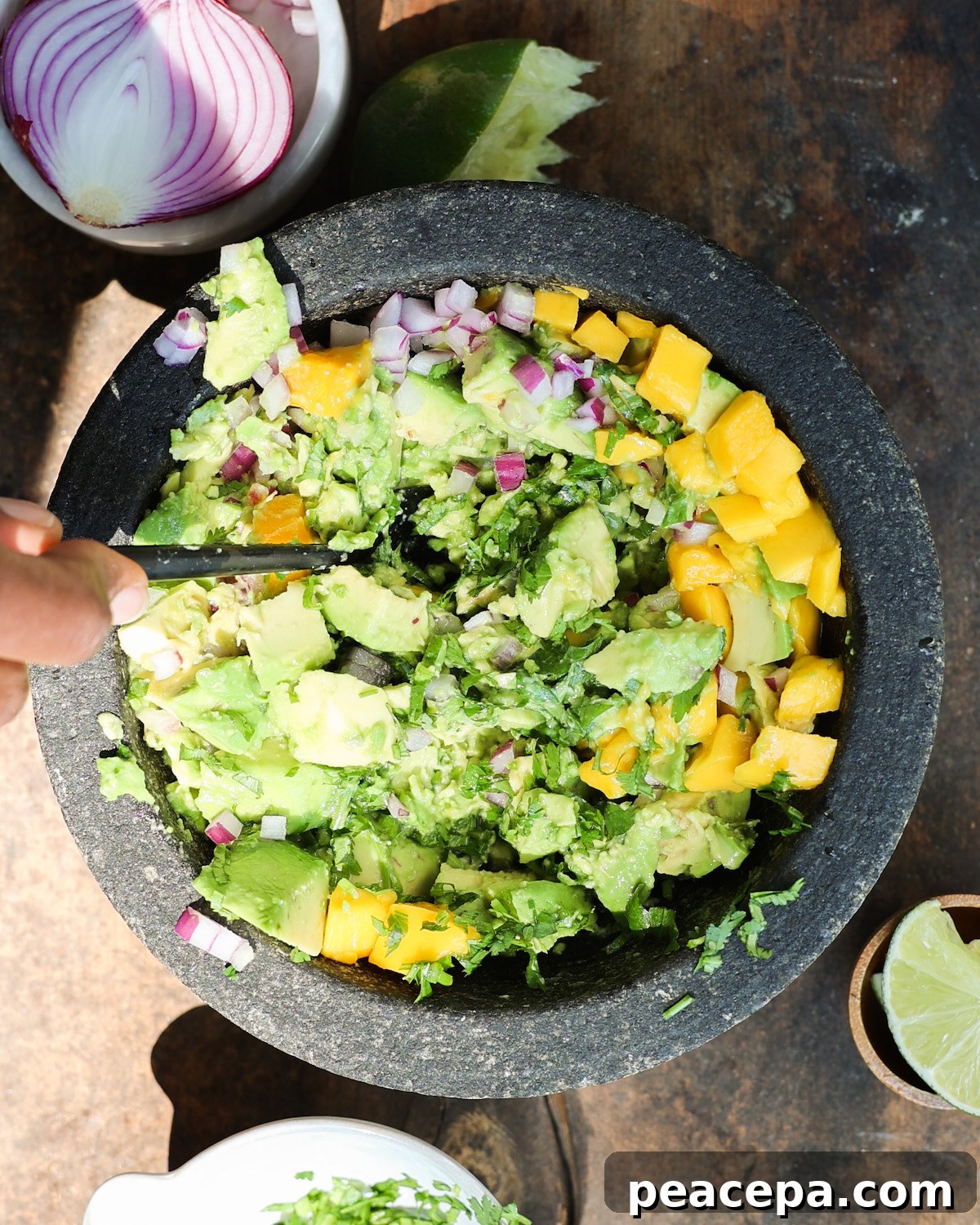 An overhead shot of a brown hand mashing the avocado, onion, herbs, and mango together.