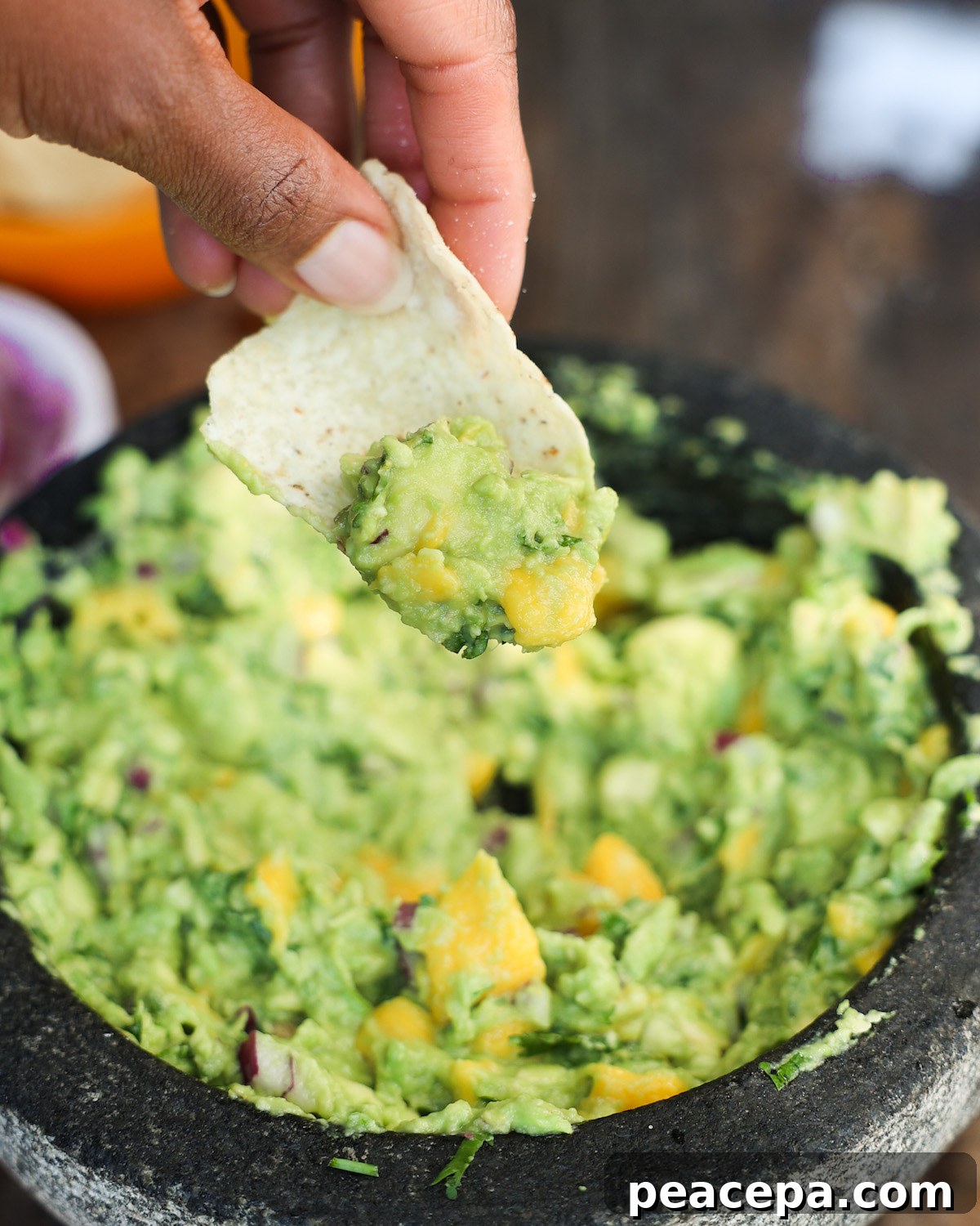 A closeup show of a brown hand holding a chip that has been dipped in mango guacamole.