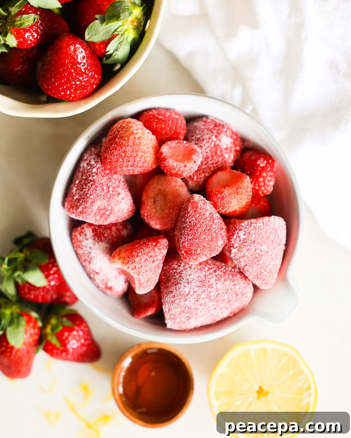 An overhead shot of a bowl of frozen strawberries next to a small cup of agave syrup, fresh strawberries, and half a lemon.