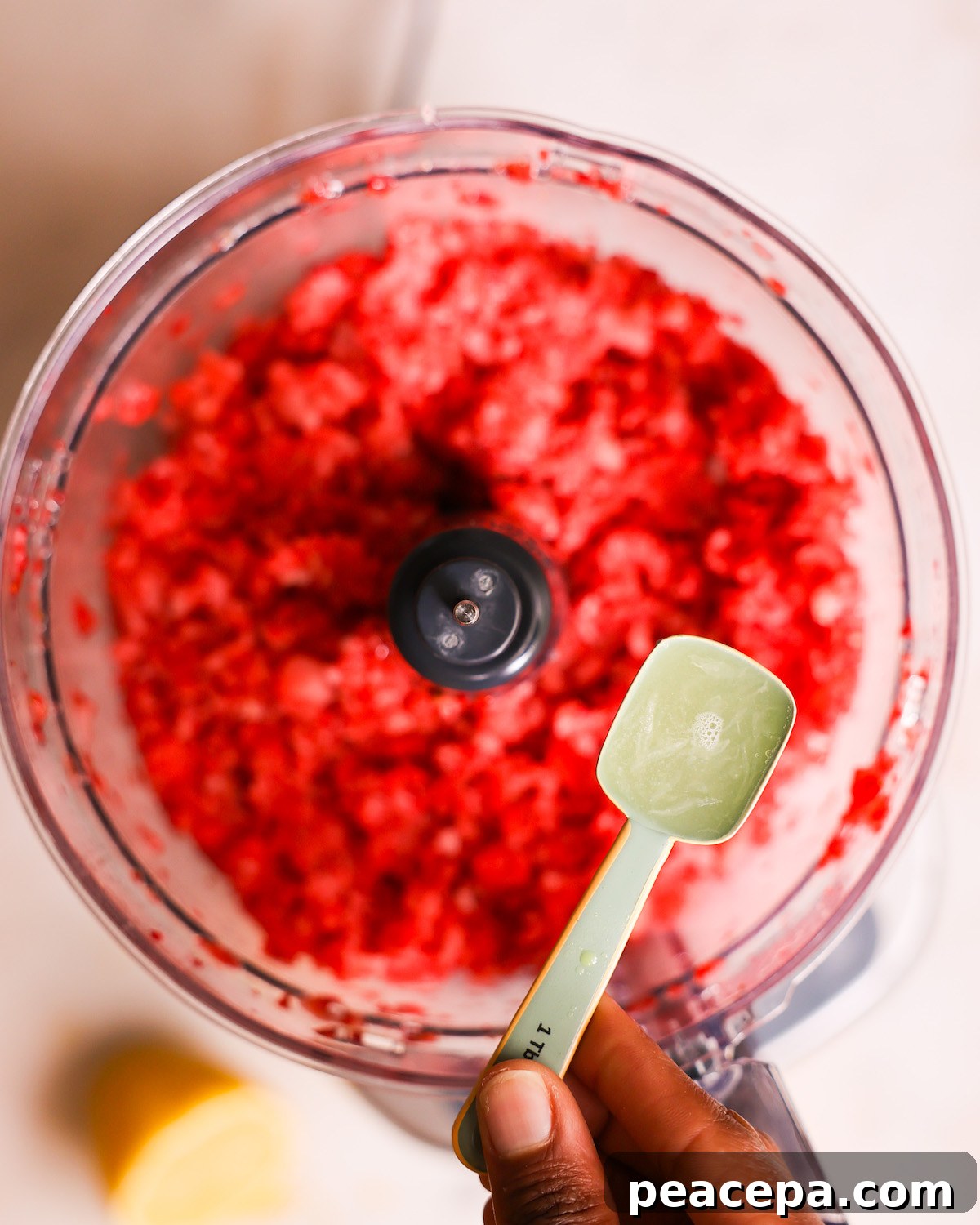 A brown hand holding a tablespoon of lemon juice over a food processor of blended strawberries.