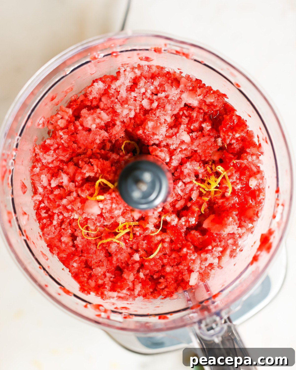 An overhead shot of a food processor with pulsed strawberries and lemon zest.
