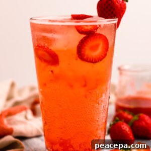 A side shot of a tall glass of strawberry soda on the counter with strawberries and a dish towel.