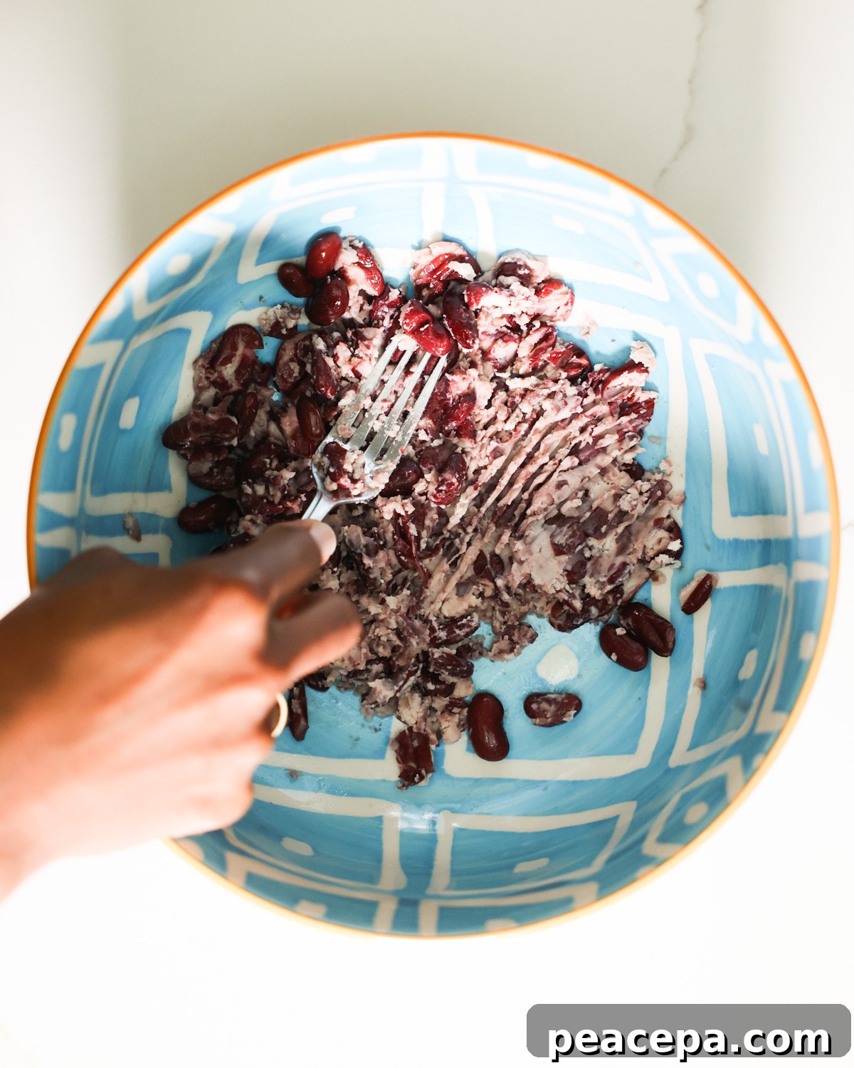 A brown hand mashing the beans in a blue patterned mixing bowl.