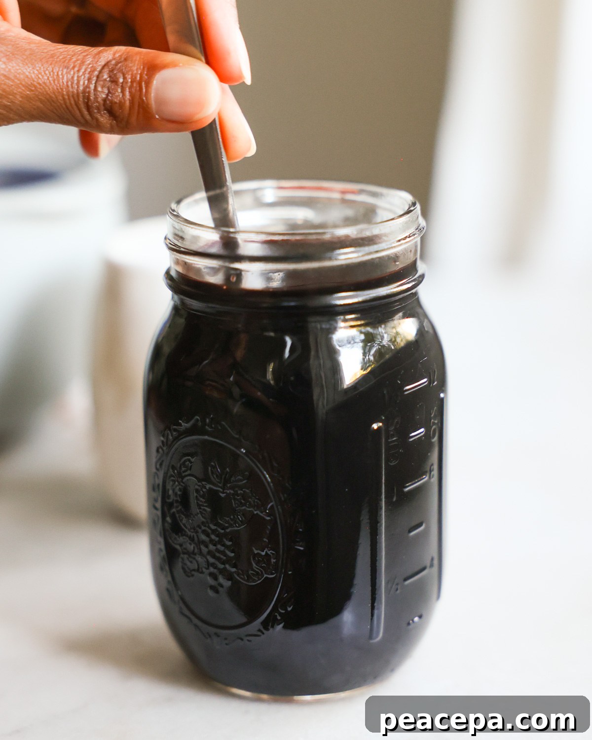 A close-up shot of a brown hand hand stirring a small har of elderberry syrup.