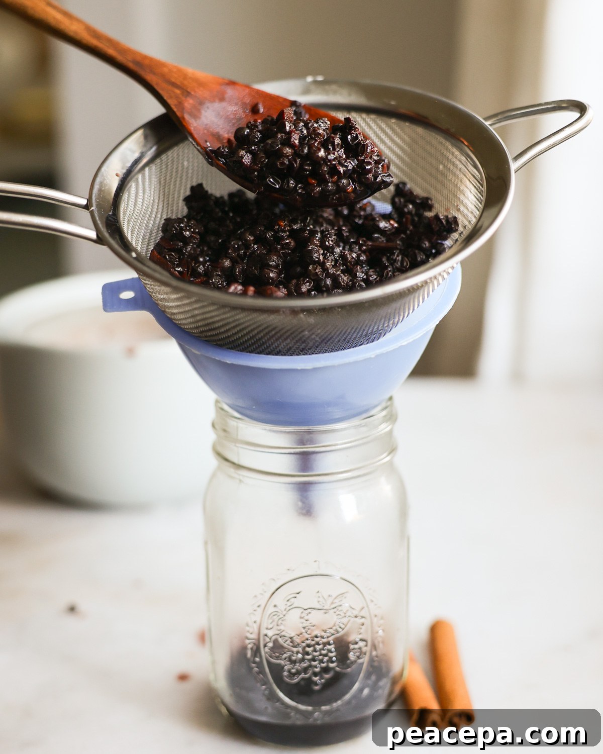 Pressing the simmered elderberry syrup through a sieve over a funnel into a glass jar.