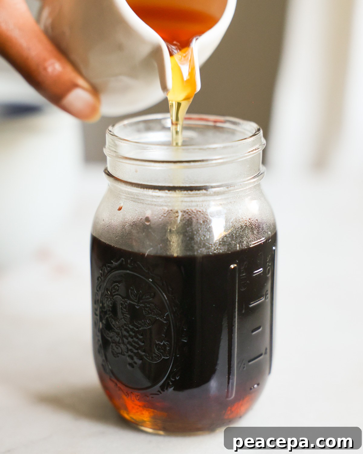 An up-close shot of a brown hand pouring agave syrup into a jar with the elderberry juice.