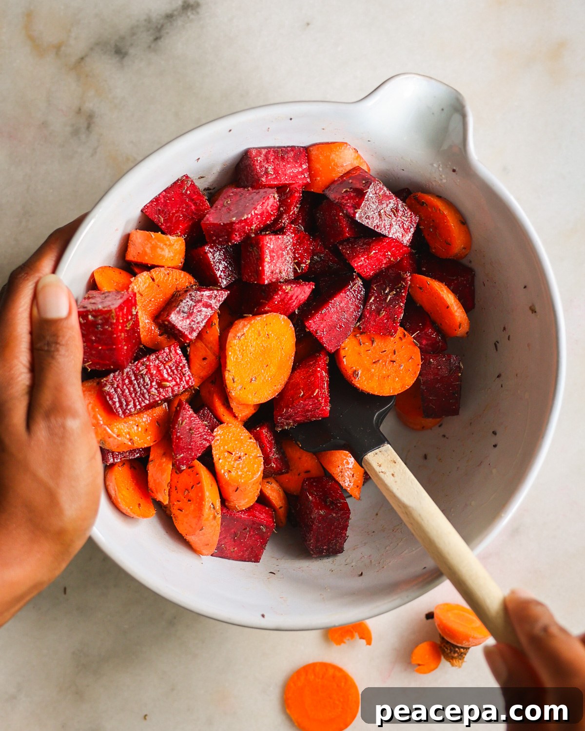 An overhead shot of a brown hand stirring the carrots and beets with seasonings in a mixing bowl.