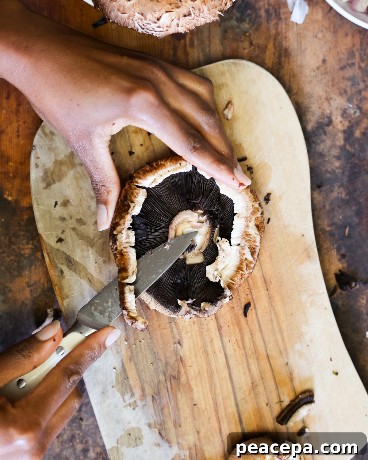 Two brown hands trimming a portobello mushroom on a small cutting board.