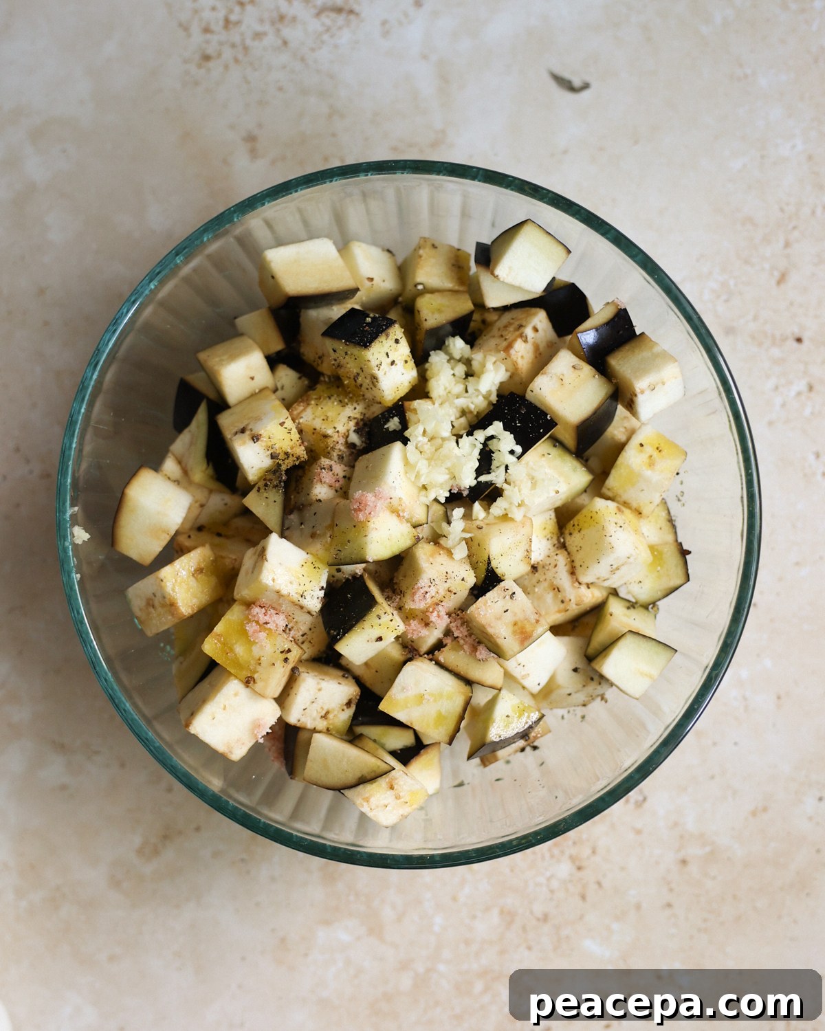 An overhead shot of a bowl of cubed eggplant topped with chopped garlic, olive oil, salt, and pepper.
