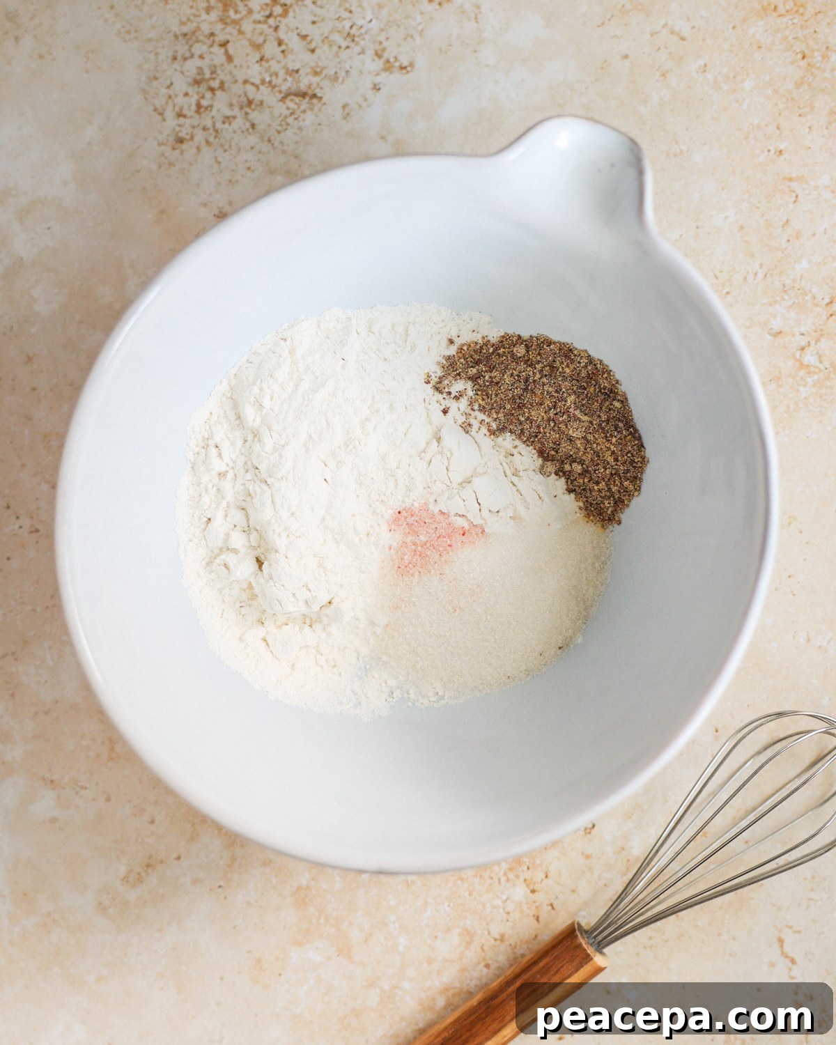 An overhead shot of the dry ingredients added to a large mixing bowl next to a whisk.