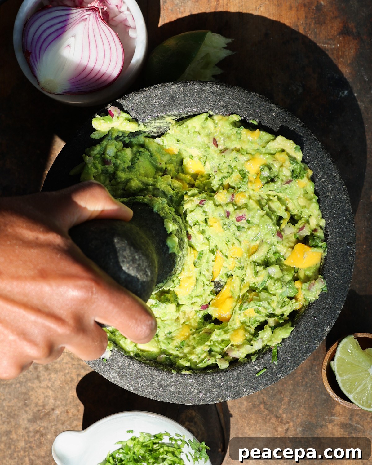 An overhead shot of a brown hand mashing the mango avocado guacamole with a mortar and pestle.