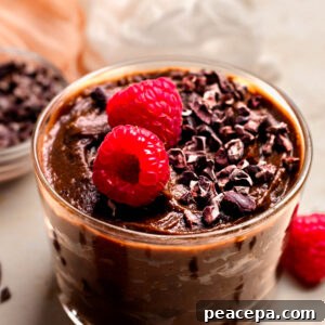 An up-close angled shot of a glass dish of vegan chocolate mousse on the counter with carob chips and raspberries.