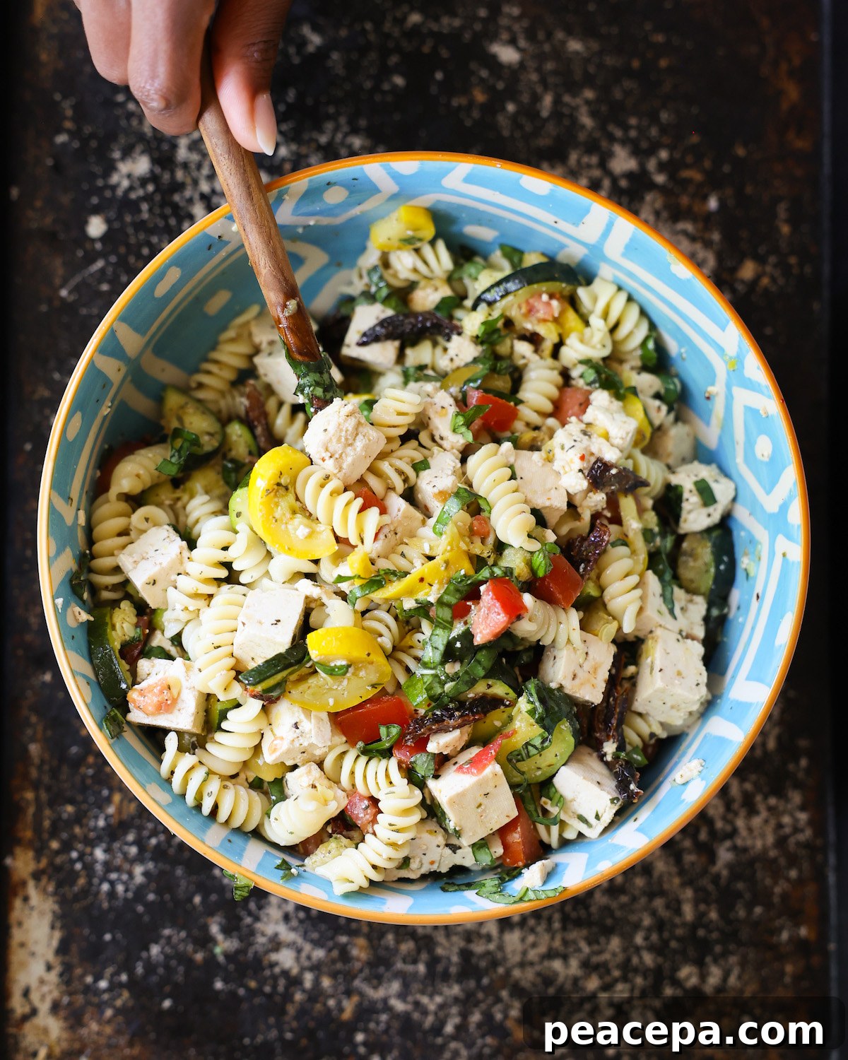An overhead shot of a brown hand stirring a bowl of Mediterranean pasta salad.