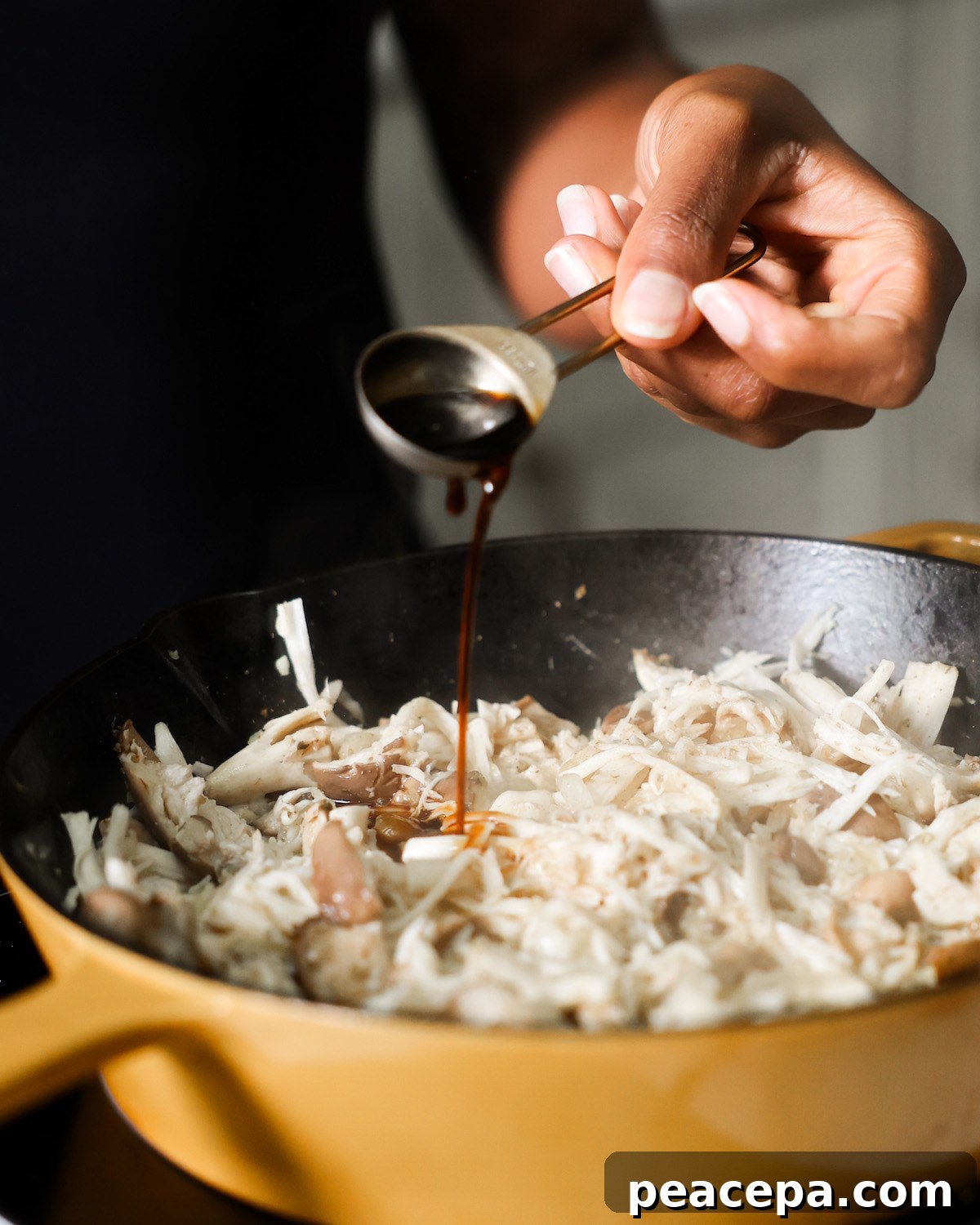 brown hand pouring soy sauce from a measuring spoon into skillet