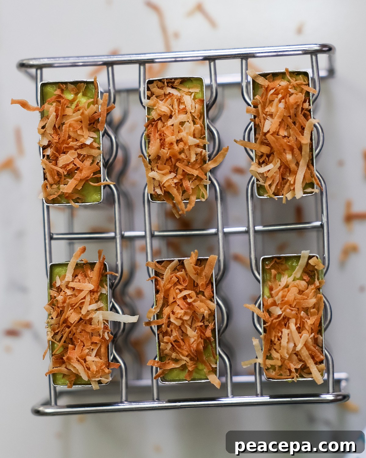 An overhead shot of metal popsicle molds filled with avocado pineapple mixture and topped with dried coconut.