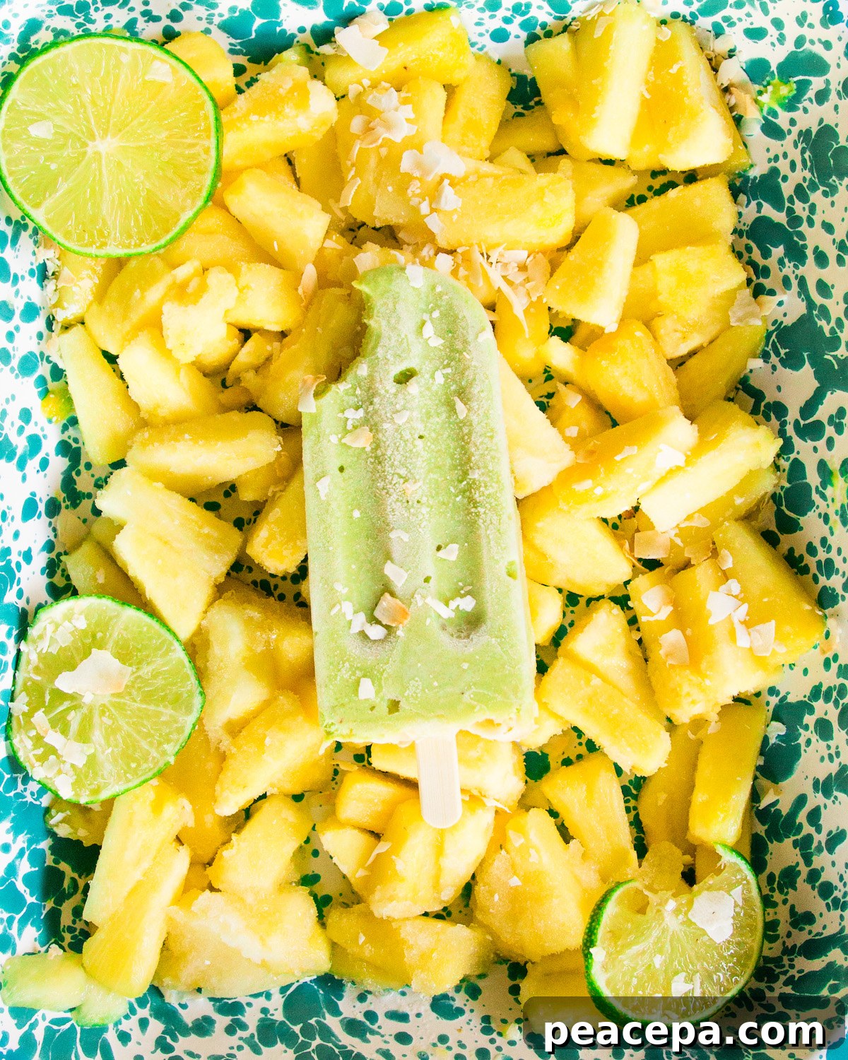 An overhead shot of an avocado pineapple popsicle with a bite taken out of it laying on a blue tray of frozen pineapple.