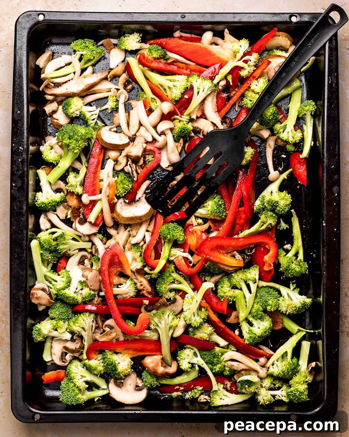 An overhead shot of a baking sheet of sliced vegetables with a black spatula.