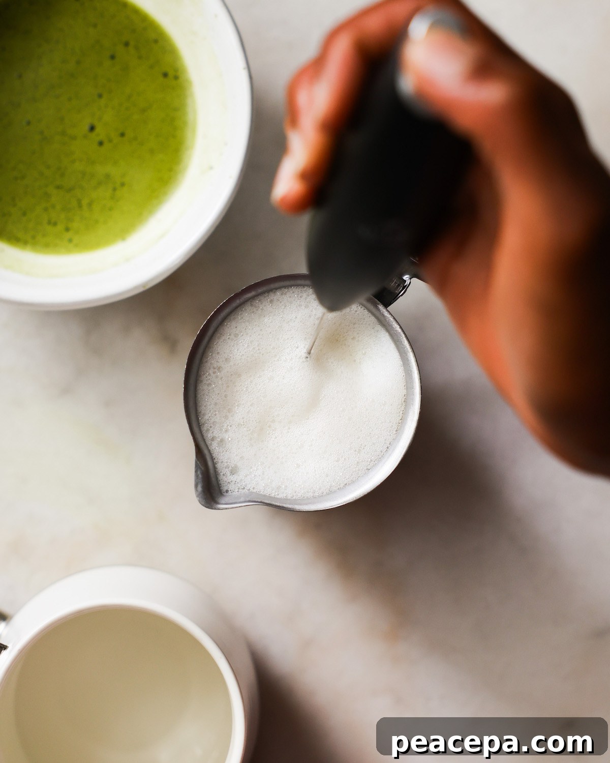 An overhead shot of a brown hand frothing a small pitcher of oat milk.