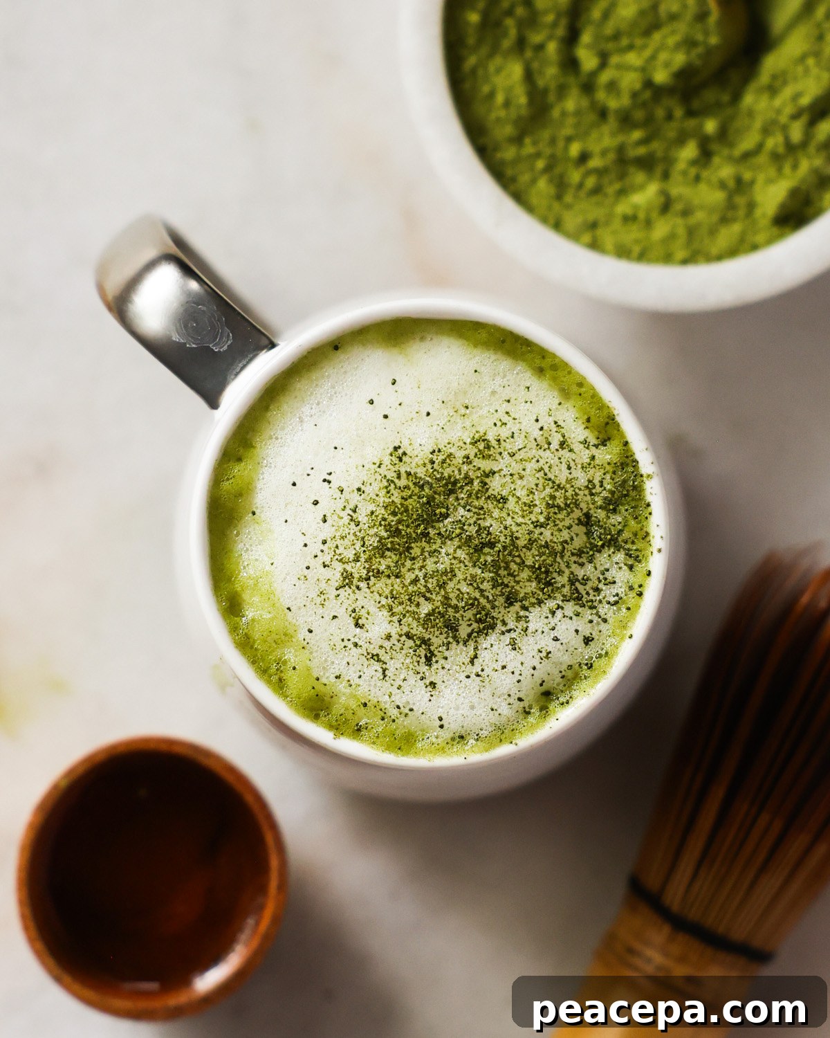 An overhead shot of a mug of the oat milk matcha latte on the counter next to a small dish of matcha and the matcha whisk.